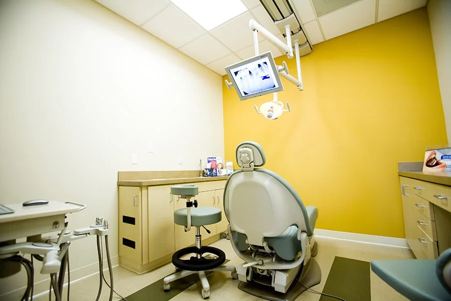 Single dental chair in exam room with yellow wall and overhead screen.