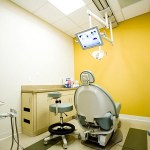 Single dental chair in exam room with yellow wall and overhead screen.